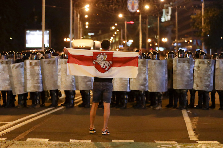 Protestor stands, holding Belarusian flag, in front of riot police.